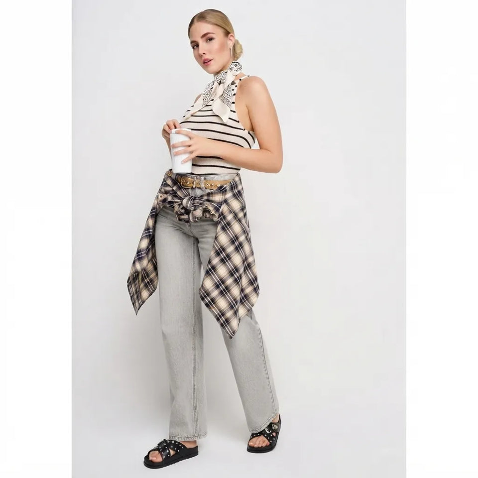 A woman models a sleeveless black and white striped top with a patterned neck scarf, light gray wide-leg jeans, and a beige plaid shirt tied around her waist, holding a white coffee cup against a light gray studio background.