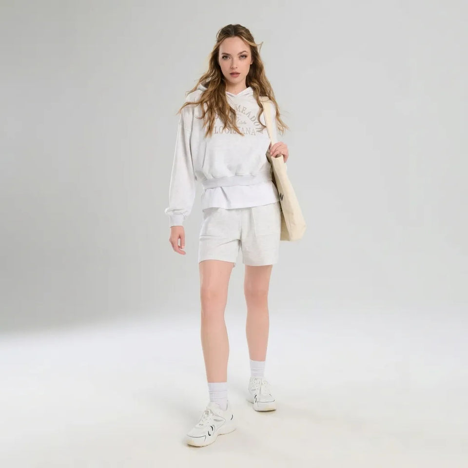 A woman modeling a white long-sleeve sweatshirt with embroidered details, matching white shorts, white socks, and white sneakers, holding a beige tote bag against a light gray studio background.