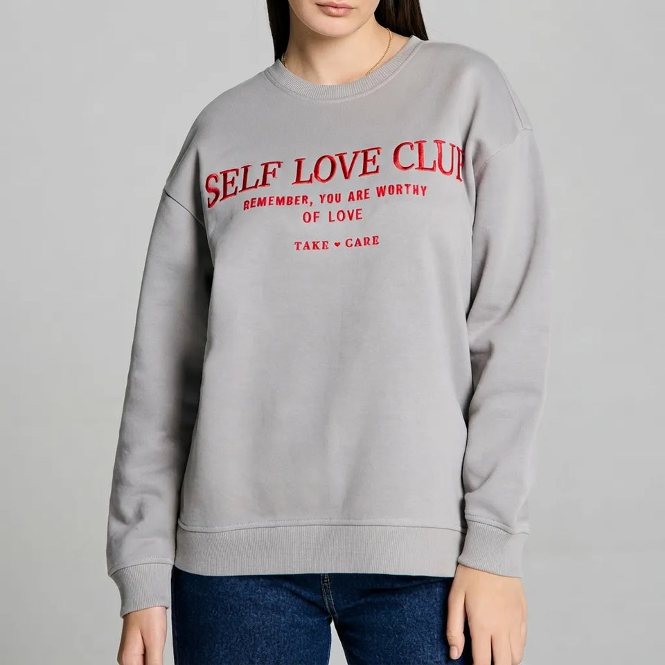 A woman wearing a light gray crewneck sweatshirt with red text that reads 'SELF LOVE CLUB' and smaller supportive phrases underneath, paired with blue jeans against a plain light gray studio background.