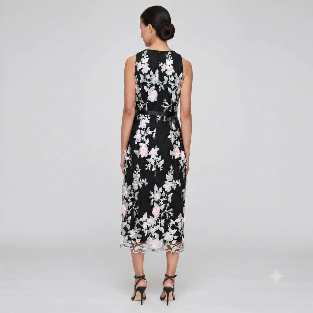 A woman stands with her back to the camera wearing a sleeveless black midi dress with a white and pink floral pattern and lace trim at the hem, paired with black strappy heels, against a light gray studio background.