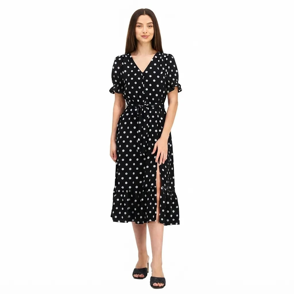 A woman modeling a black midi wrap dress with white polka dots, short puff sleeves, and a front slit, paired with black slide sandals against a plain white background.