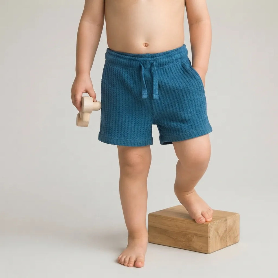 A child wearing textured teal blue shorts stands barefoot on a wooden block against a light gray studio background, holding a small wooden toy.