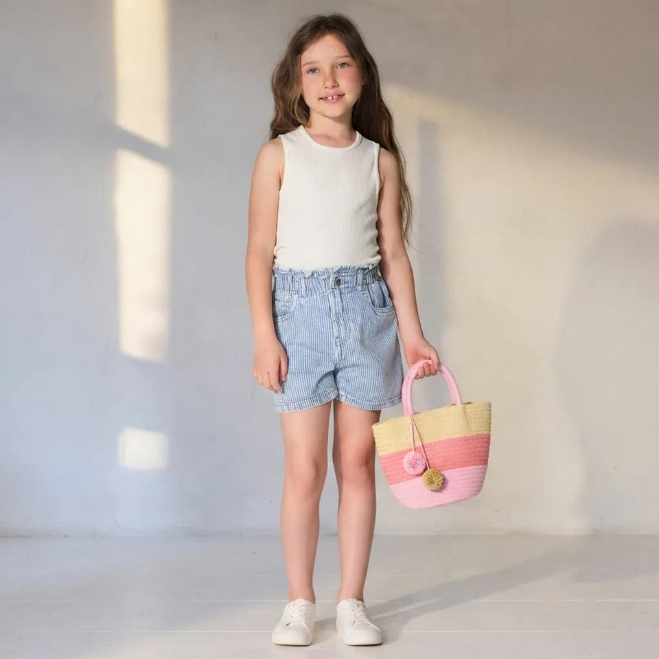 A young girl stands in a bright studio wearing a cream sleeveless top, light blue denim shorts, and white sneakers, holding a pastel striped woven tote bag.