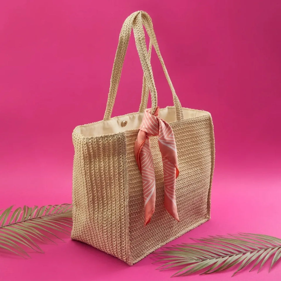 A woven straw tote bag with dual shoulder straps, featuring a coral pink patterned silk scarf tied around the handle, set against a vibrant pink background with decorative palm leaves at the bottom.