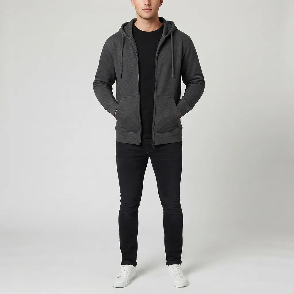 A man wearing a dark gray zip-up hoodie over a black t-shirt, black jeans, and white sneakers, standing against a light gray studio background.