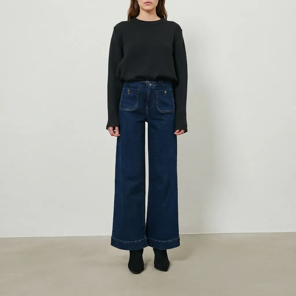 A model stands against a plain light gray studio wall wearing a black ribbed long-sleeve sweater, dark blue wide-leg denim jeans, and black ankle boots.