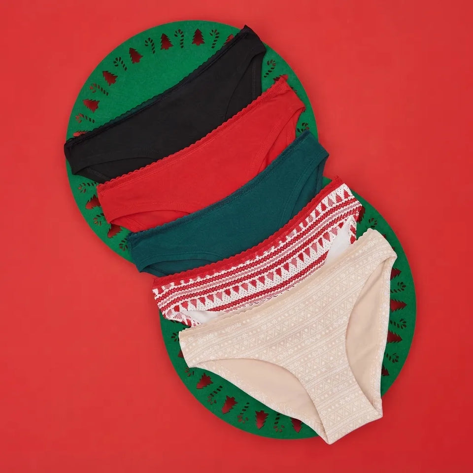 Four pairs of women's bikini-style underwear arranged in a fan shape on a green circular mat with a red background. The colors are black, red, teal, white with red pattern, and beige with white pattern.