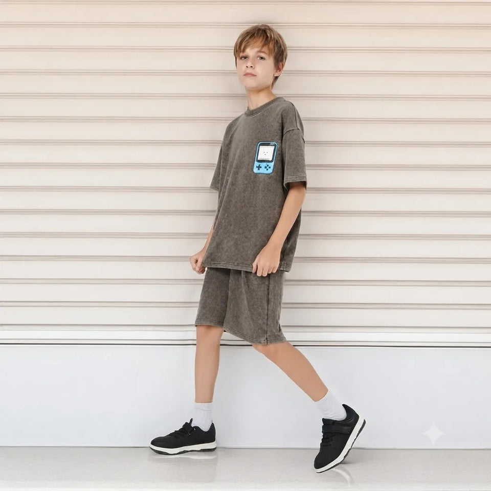 A child model wearing a matching gray short-sleeve t-shirt and shorts set with a retro handheld game console graphic on the chest, standing against a beige horizontal-slat wall.