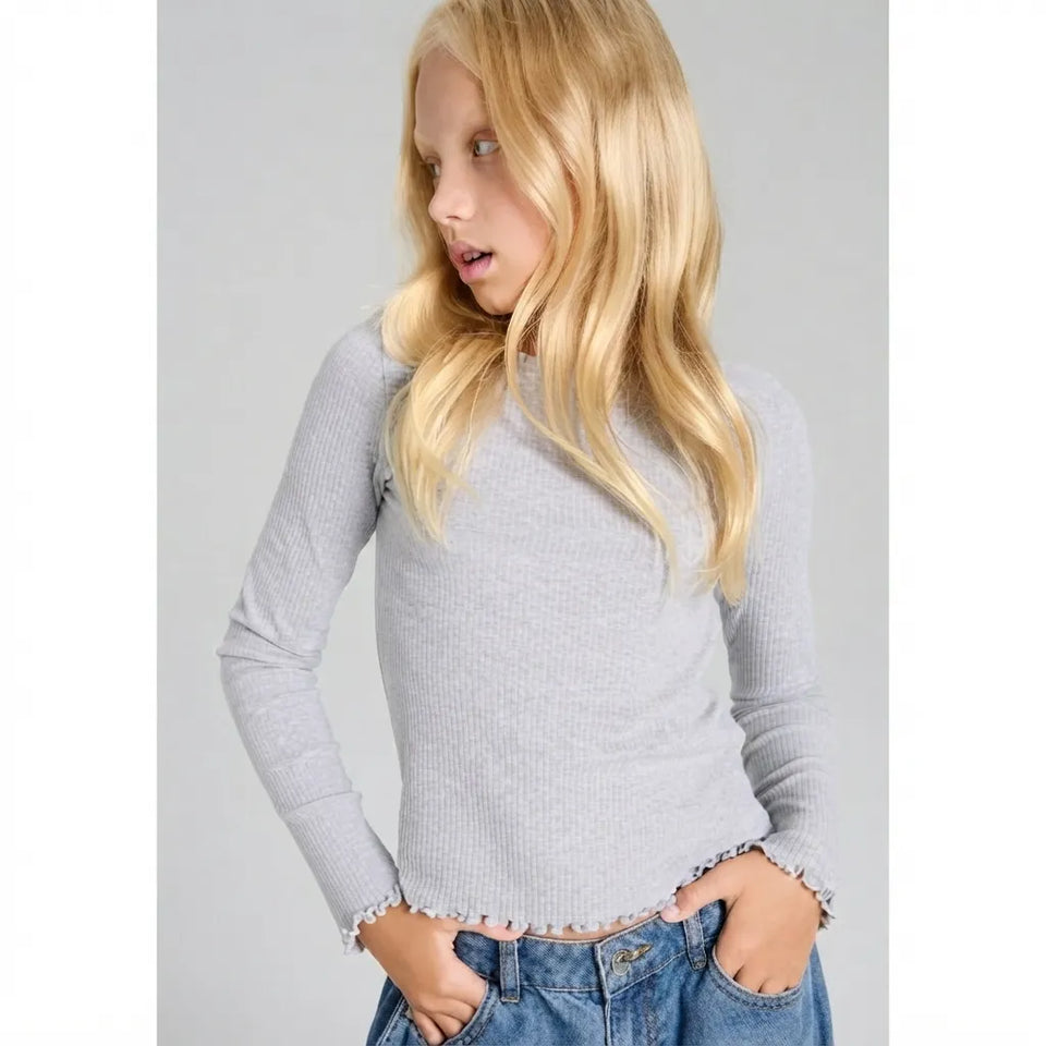 A young blonde girl wearing a light gray ribbed long-sleeve top with a raw, frayed hem and cuffs, paired with blue jeans, shown against a plain gray background.