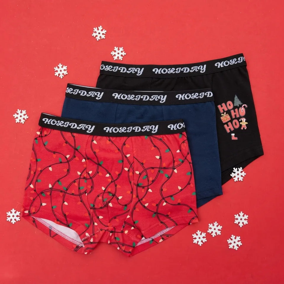 Three pairs of Rosemary Christmas-themed boxer briefs arranged in an overlapping fan on a bright red background with white snowflake decorations. The red pair features a Christmas light pattern, the navy pair is solid navy, and the black pair has a 'Ho Ho Ho' graphic.