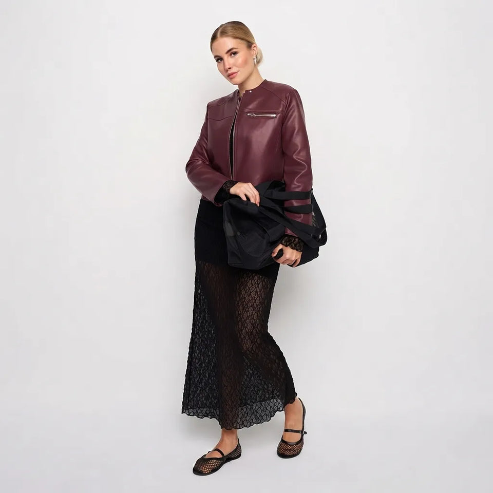 A woman models a burgundy faux-leather moto jacket with black mesh sleeves, paired with a black skirt and holding a black jacket, against a plain white studio background.