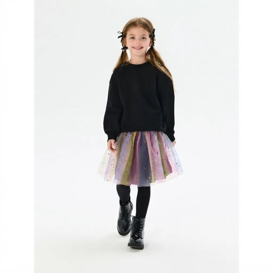 A young girl with pigtails is wearing a black long-sleeve crewneck sweater, a sparkly pastel tulle tutu skirt in purple, pink, and gold, black tights, and black boots, standing against a plain white studio background.