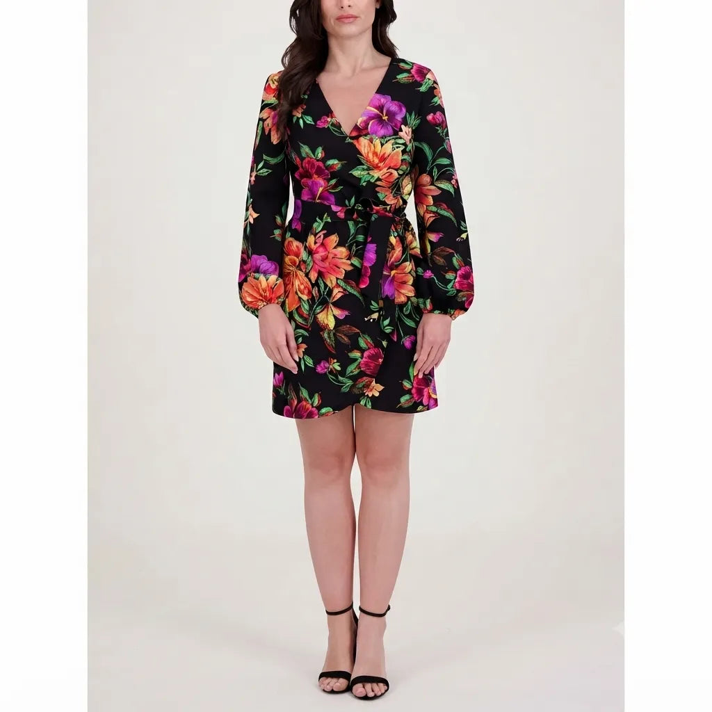 A woman models a black long-sleeve mini dress with a vibrant multicolor floral print, paired with black strappy high-heeled sandals, against a plain light beige studio background.