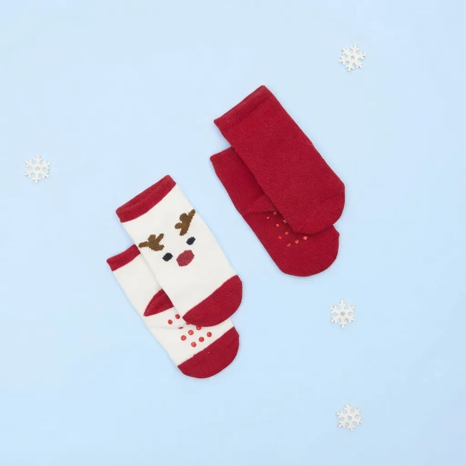 A pair of red and white Christmas-themed baby socks with a reindeer face design, laid flat on a light blue background with small white snowflake decorations.