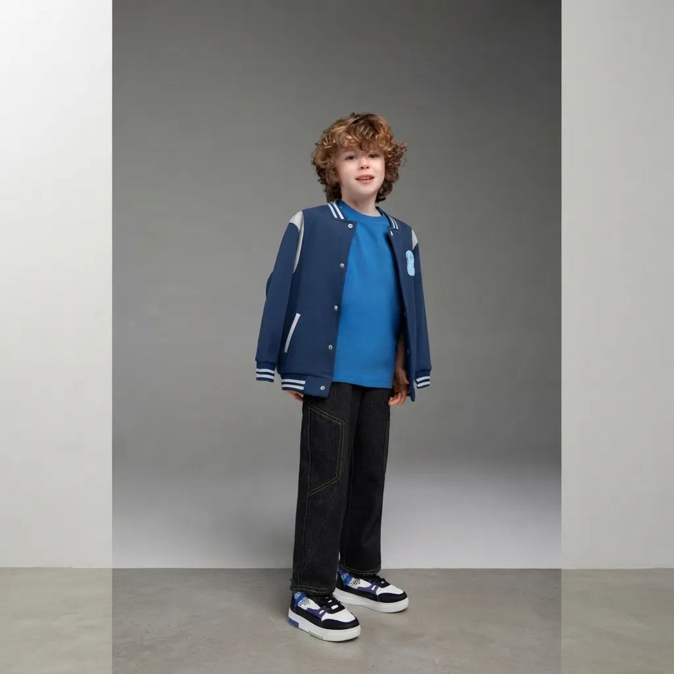 A young boy with curly brown hair stands in a studio wearing a navy blue varsity-style jacket with white stripes, a bright blue t-shirt, dark jeans, and navy sneakers.