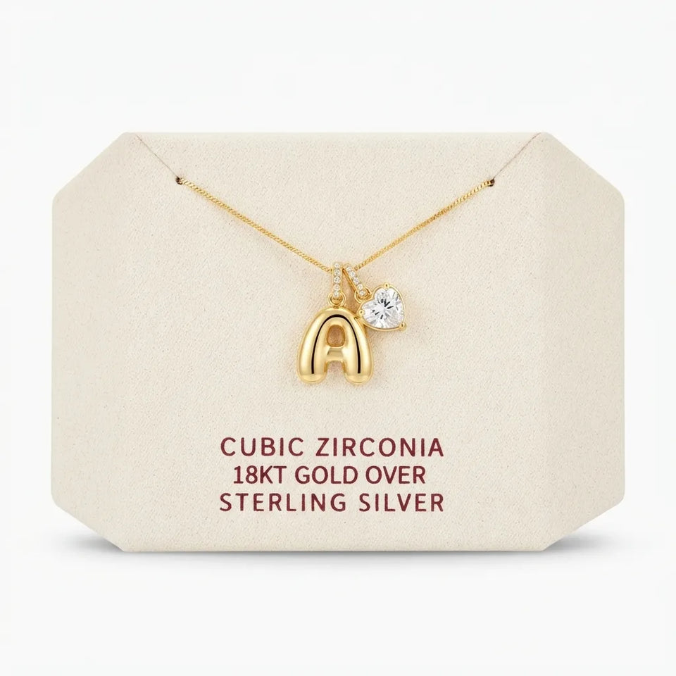 A gold necklace with a letter 'A' pendant and a small heart-shaped cubic zirconia charm, displayed on a cream-colored jewelry card with text detailing the materials.