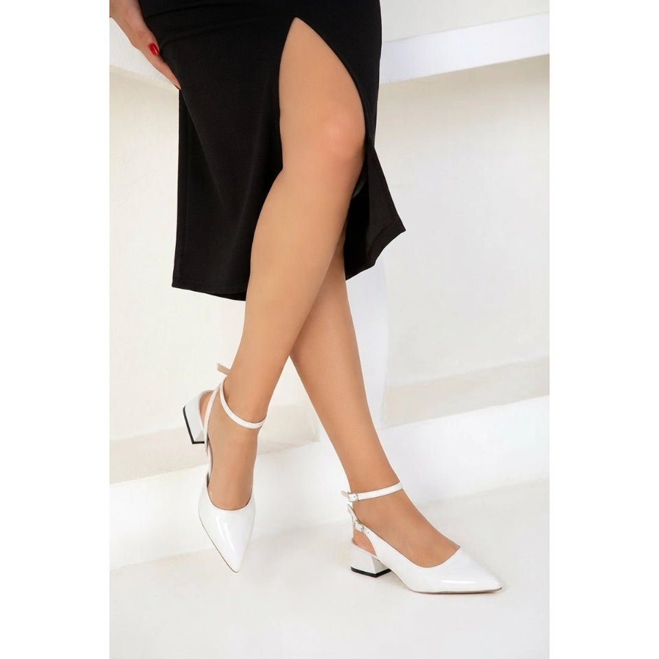 White pointed-toe slingback heels with ankle straps and low block heels, shown on a model wearing a black skirt in a bright white interior setting.