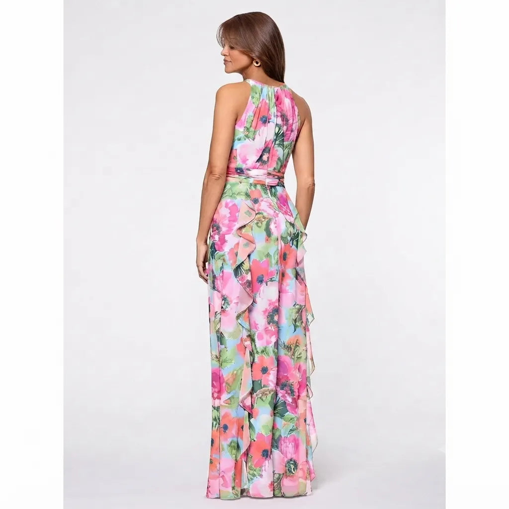 A woman modeling a sleeveless halter-neck maxi dress with a vibrant pink, green, and blue floral print, shown from a rear three-quarter view against a light gray studio background.