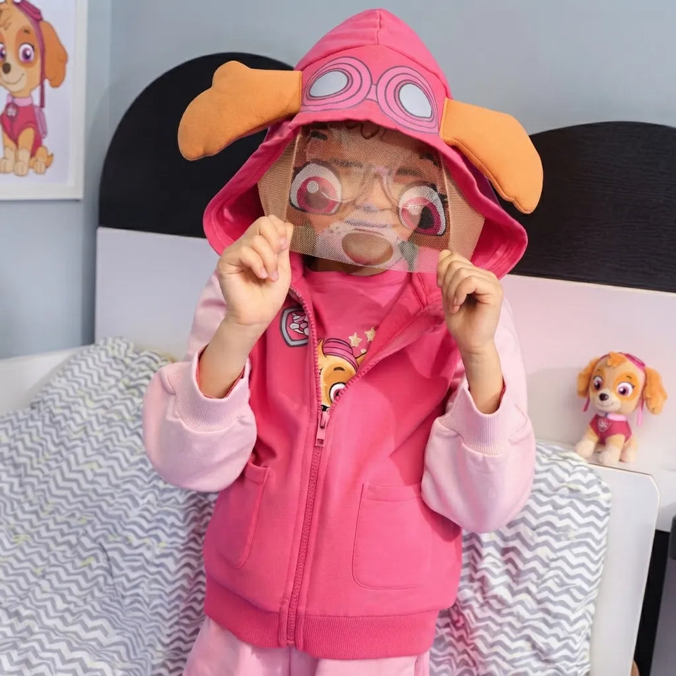 A child wearing a pink Paw Patrol zip-up hoodie with a hood featuring dog ears and goggles, holding a Paw Patrol-themed visor in front of their face, in a bedroom setting.