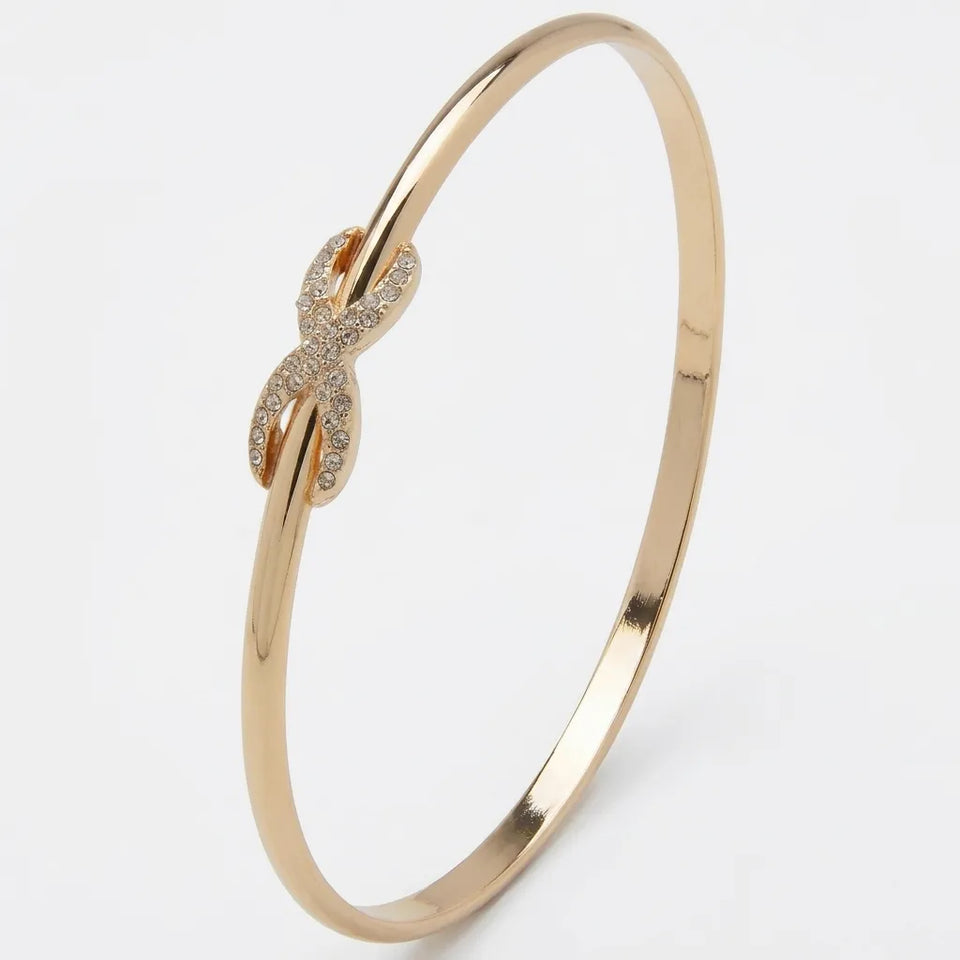 A polished gold bangle bracelet featuring a central infinity symbol paved with small clear stones, set against a clean white background.