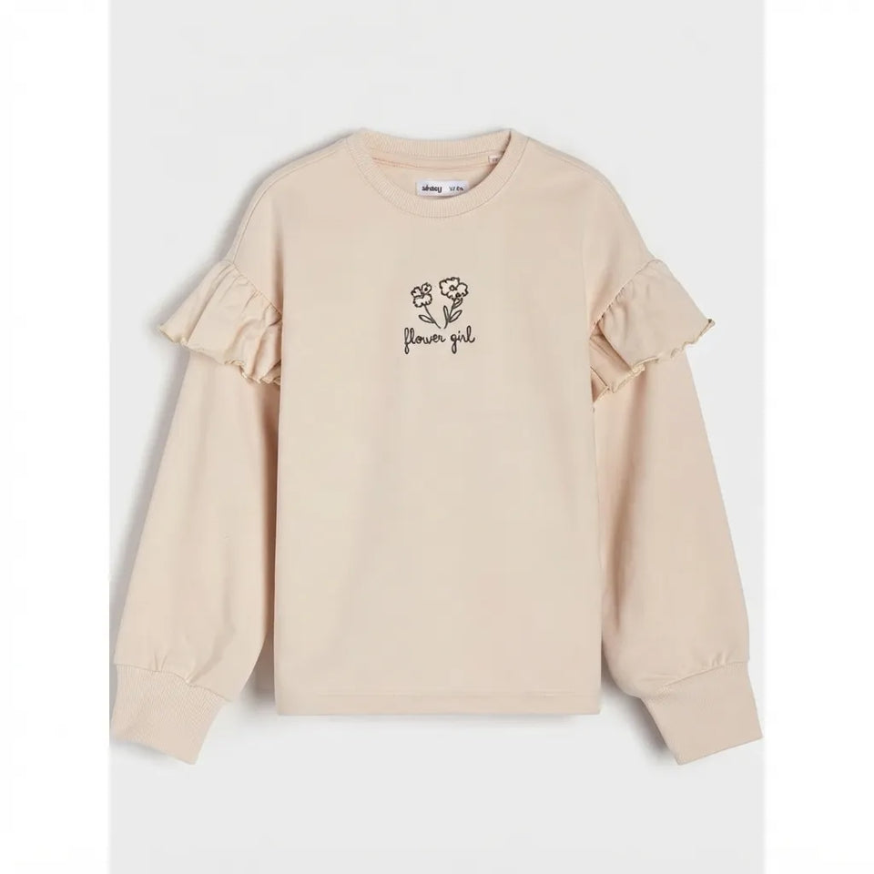 A light beige long-sleeve sweatshirt with ruffled details on the shoulders and sleeves, featuring a small black embroidered graphic of flowers and the text 'flower girl' on the chest, displayed against a light gray background.