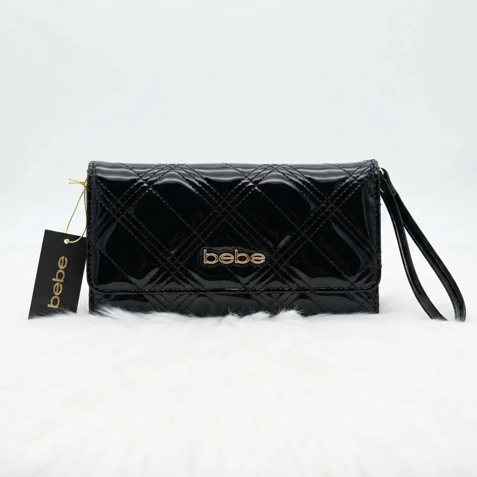 A black patent leather clutch bag with a quilted diamond pattern, featuring a gold-tone 'bebe' logo on the front and a matching black hangtag, displayed on a white faux fur surface.