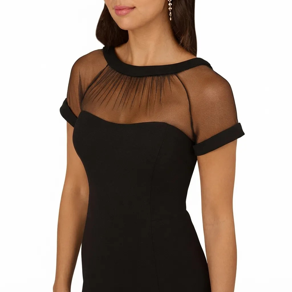 A woman wearing a black off-the-shoulder dress with sheer mesh shoulder panels and a structured neckline, shown against a plain white background.