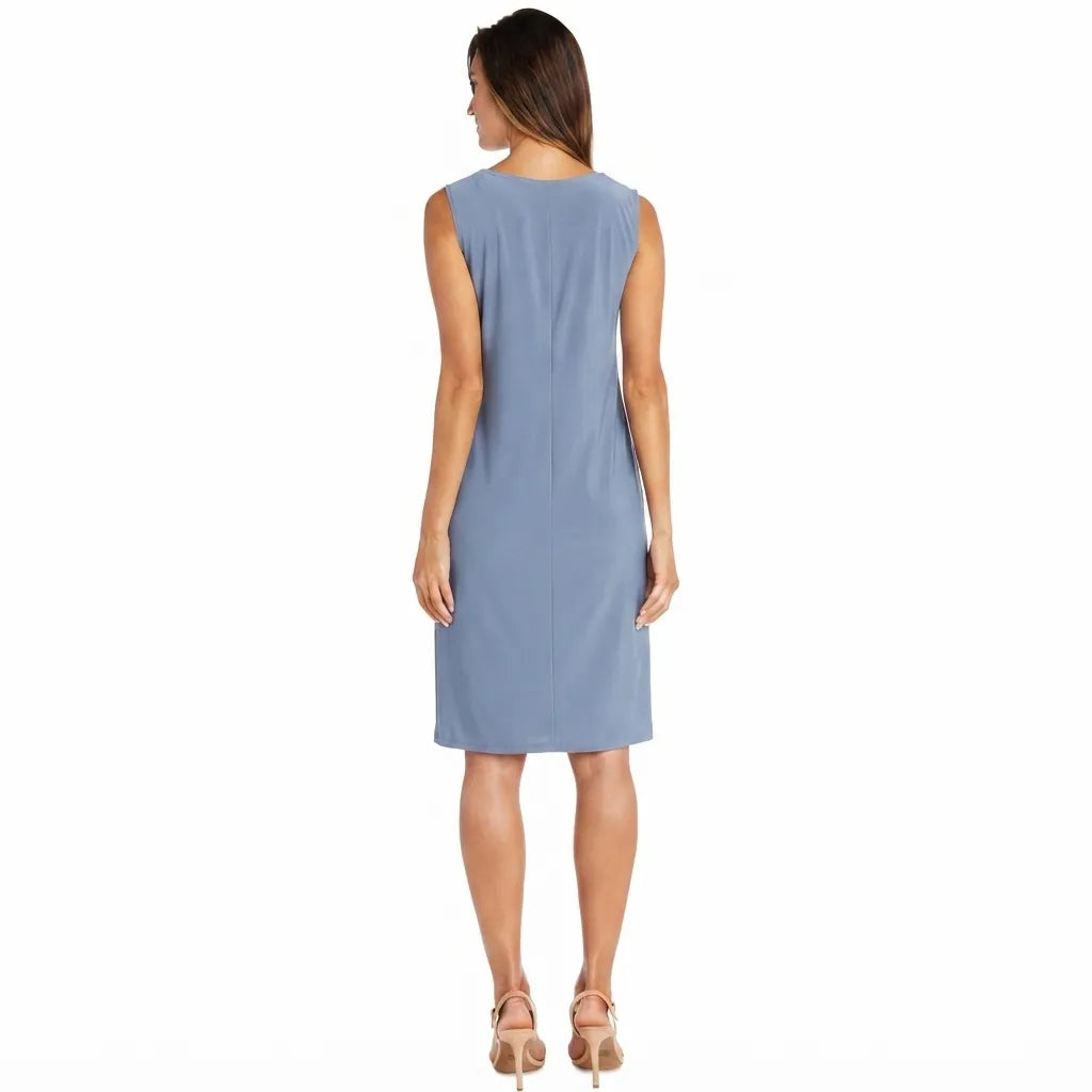A woman viewed from the back wearing a sleeveless knee-length dress in a dusty light blue color, paired with beige high-heeled sandals, against a plain white background.