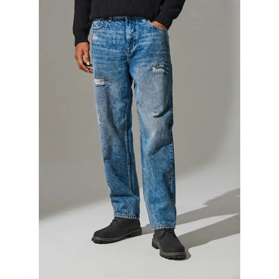 A person wearing medium-wash blue distressed jeans with faded details, a black sweater, and black shoes, standing against a light gray studio background.