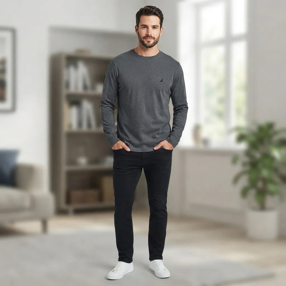 A man wearing a dark gray long-sleeve crewneck shirt with a small black logo on the left chest, paired with black pants and white sneakers, standing in a bright, modern living room.