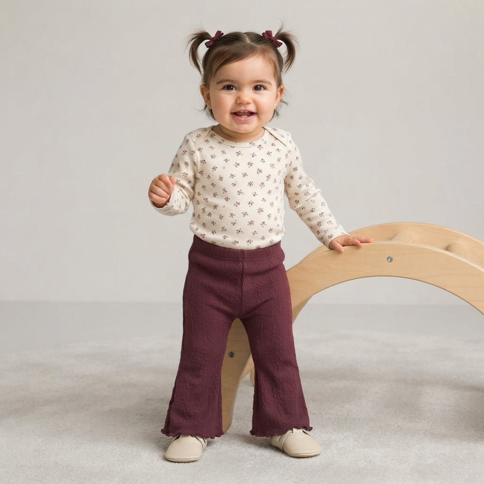 A toddler girl standing on a light gray carpet next to a wooden arch, wearing a cream long-sleeve top with a tiny floral pattern and dark burgundy flared pants.