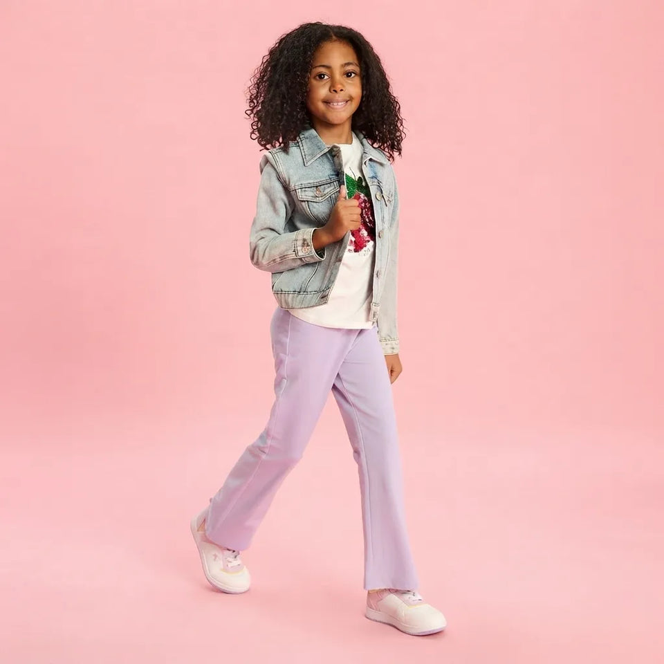 A young girl with curly black hair is modeling a light-wash denim jacket, a white t-shirt with a red cherry graphic, lavender flare-leg pants, and white sneakers, set against a solid pink studio background.