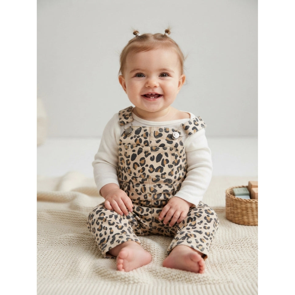 A smiling baby girl sitting on a cream knit blanket, wearing a beige and black leopard print overall romper over a white long-sleeve shirt. She has two small hair bows and is posed in a bright, neutral studio setting with a small woven basket to her side.