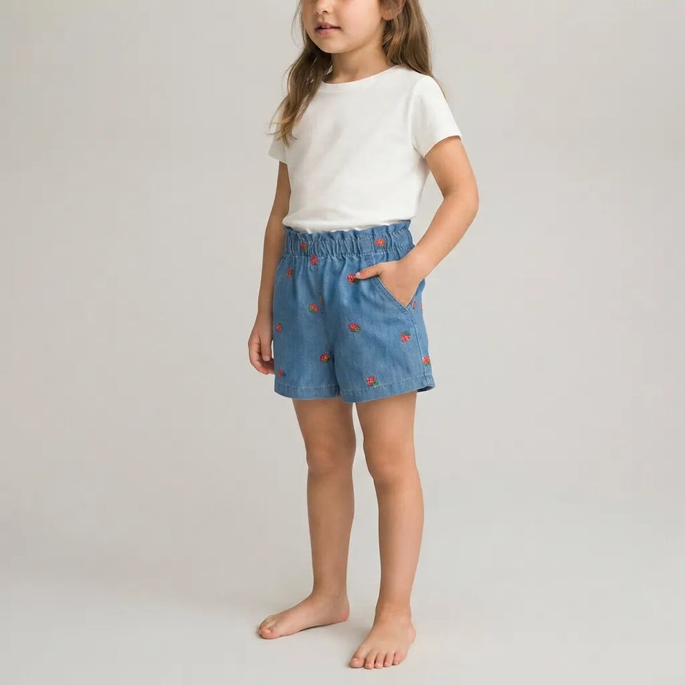 A young girl stands barefoot against a light gray studio background, wearing a white short-sleeve T-shirt and blue denim shorts with small red embroidered details.