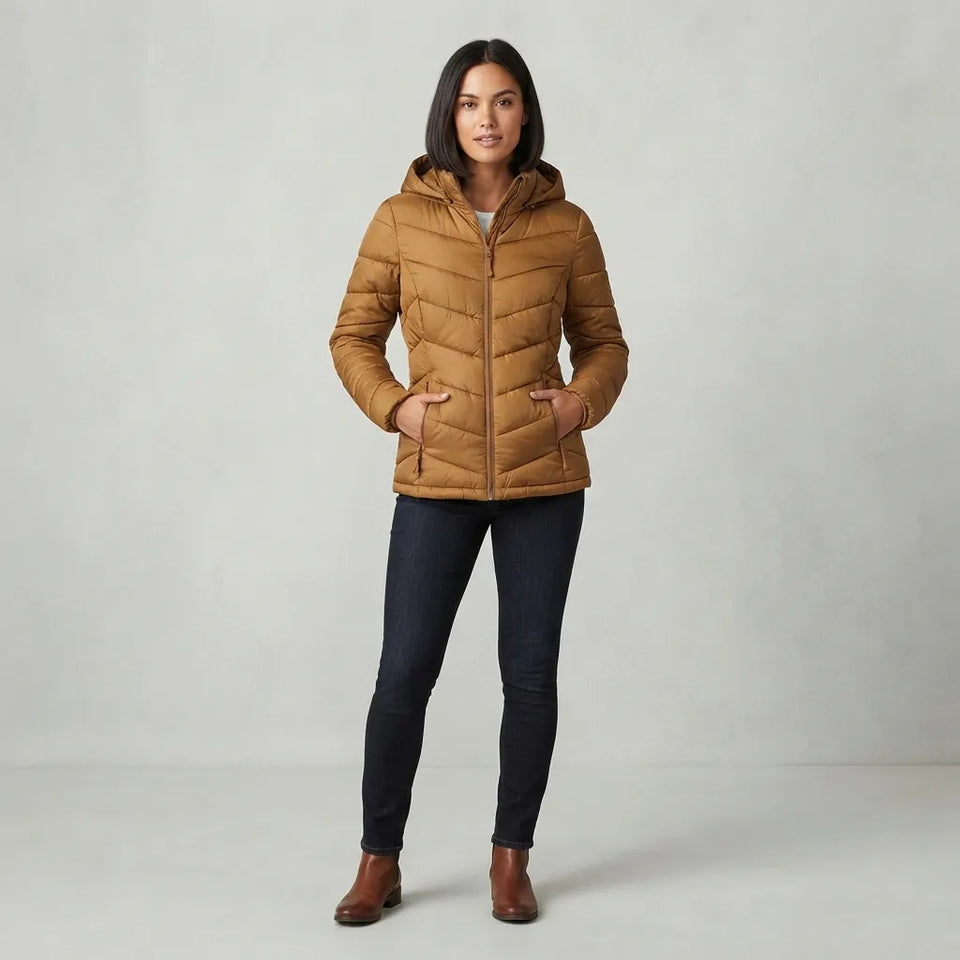 A woman modeling a tan hooded puffer jacket, paired with dark blue skinny jeans and brown ankle boots, standing against a light gray studio background.