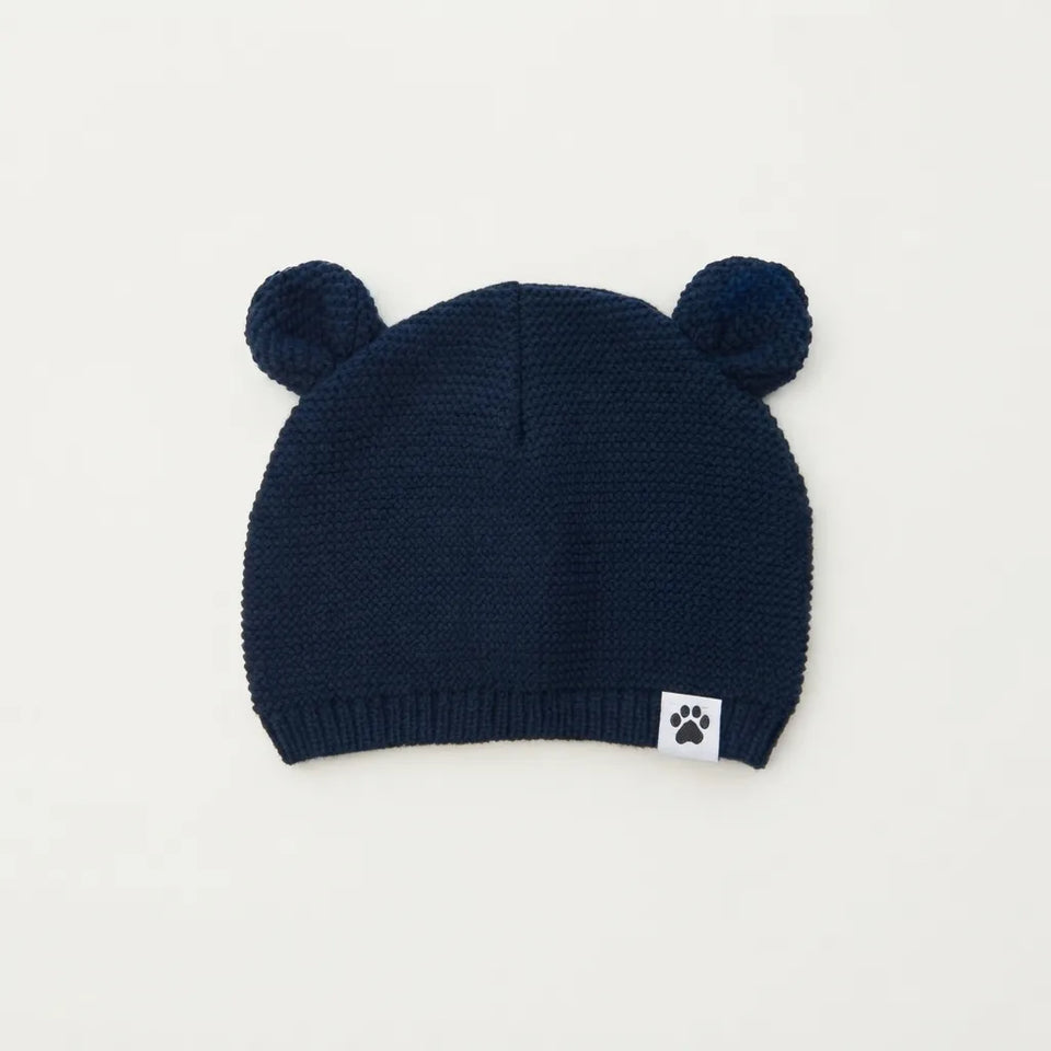 A navy blue knit baby beanie hat with two pom-pom ears on top and a small white square patch with a black paw print near the brim, shown on a plain off-white background.
