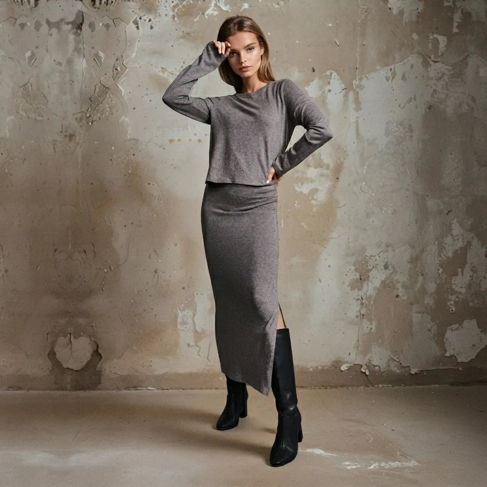 A woman models a gray knit two-piece set consisting of a long-sleeve cropped top and a matching high-slit midi skirt, paired with black knee-high boots, set against a textured concrete wall.
