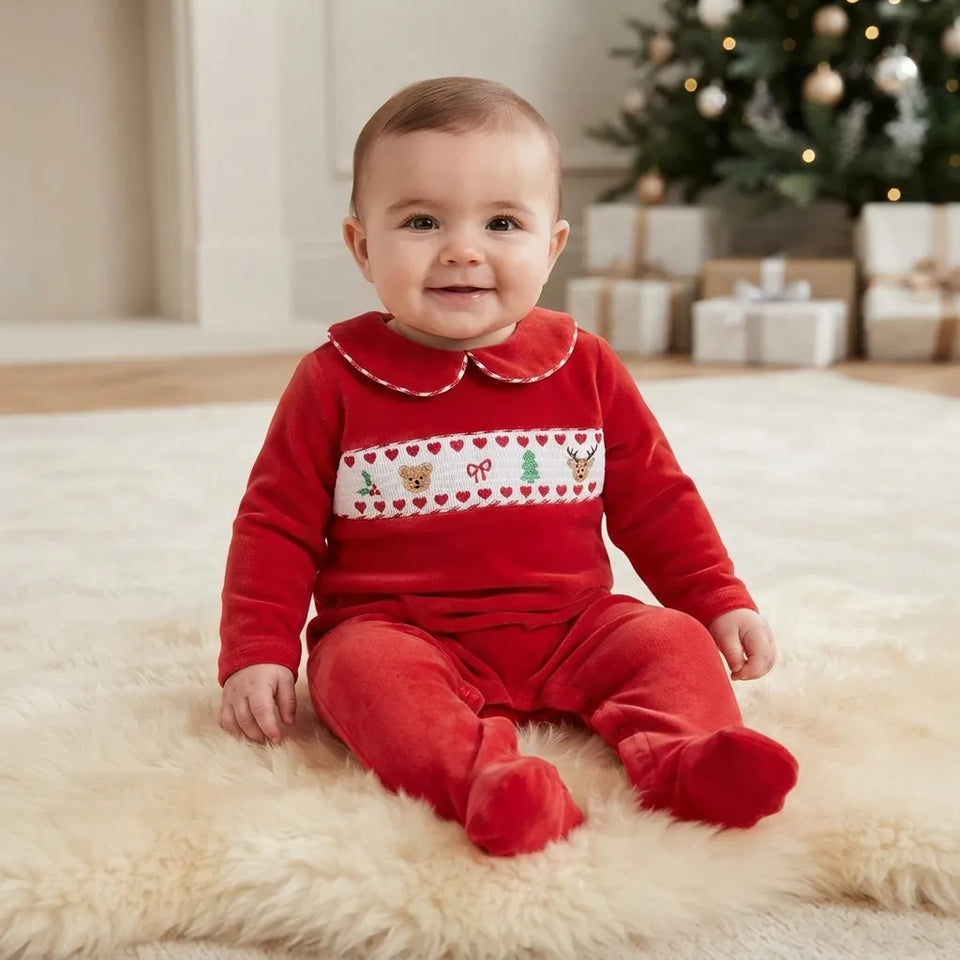A baby sitting on a fluffy cream rug wearing a red Christmas-themed footed sleeper with a white embroidered chest band featuring hearts, a Santa hat, a Christmas tree, and a bow. A decorated Christmas tree and wrapped gifts are visible in the background.