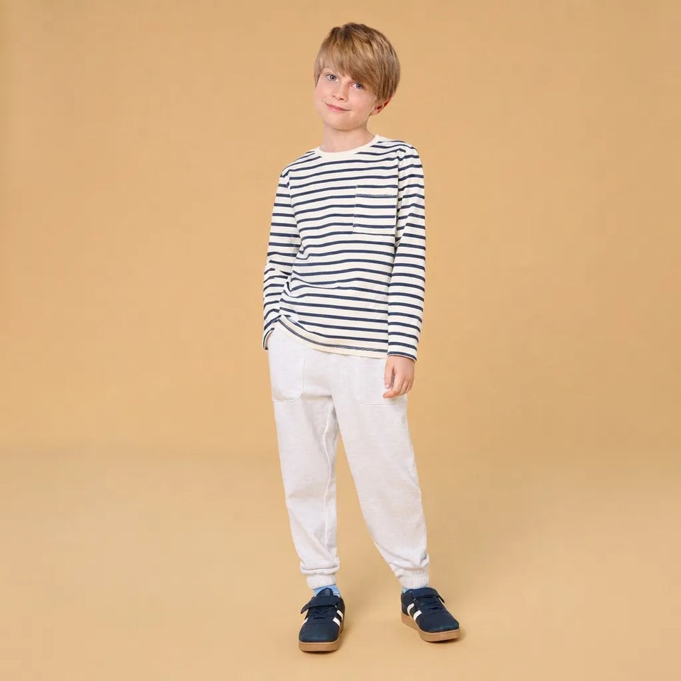 A young boy modeling a long-sleeve navy and white striped shirt, white jogger pants, and navy blue sneakers against a solid beige studio background.