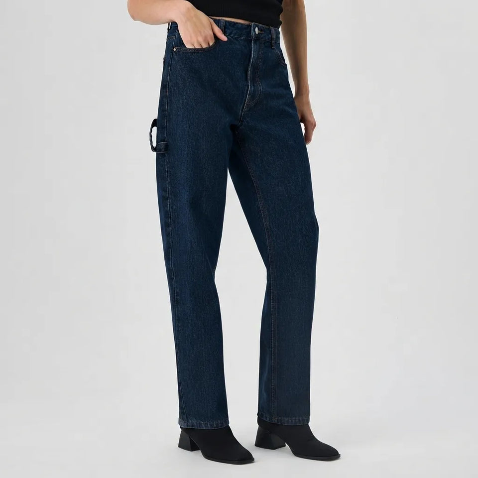 A model wearing dark blue straight-leg denim jeans with a distinctive black metal ring detail on the side pocket, paired with black ankle boots against a light gray studio background.