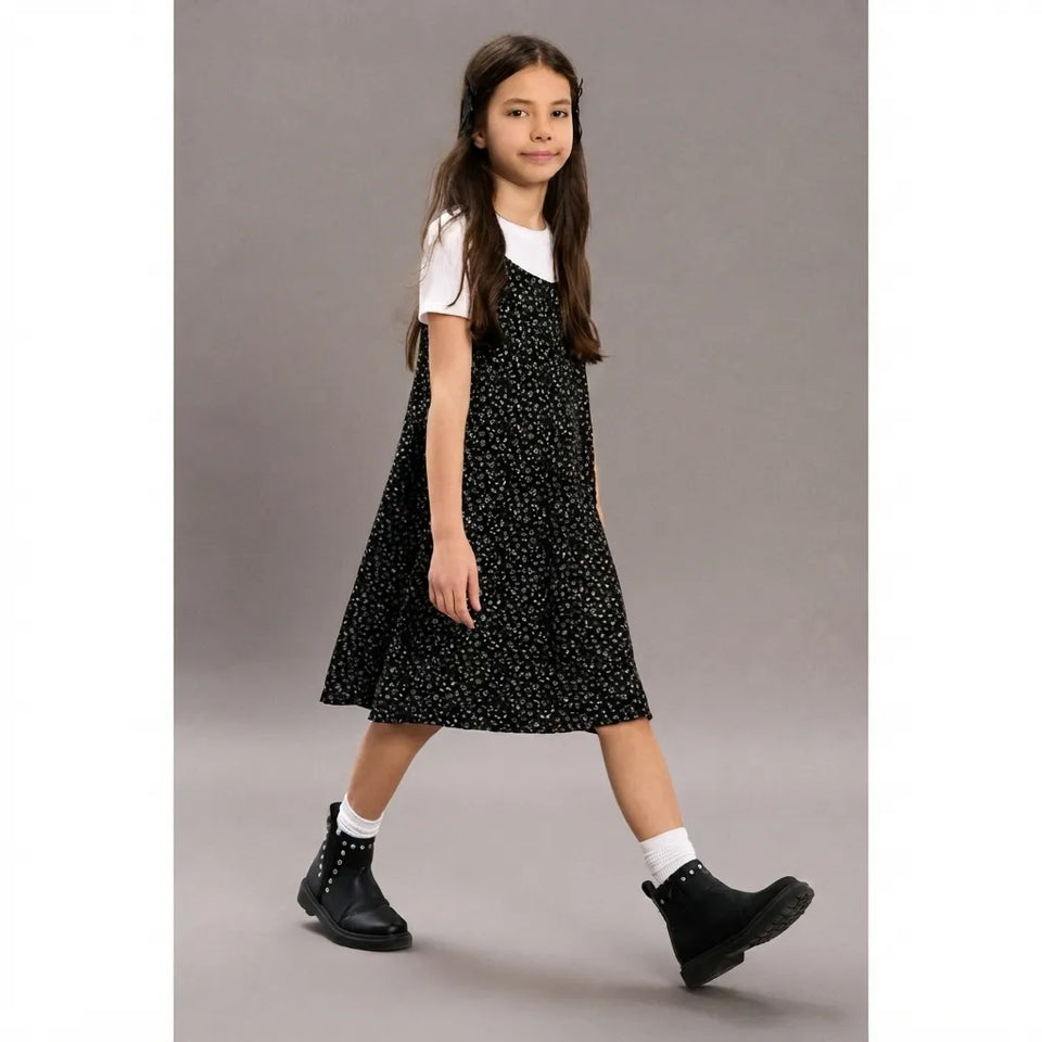 A young girl wearing a black sleeveless dress with a small white floral pattern over a white t-shirt, paired with white socks and black ankle boots, walking against a solid gray studio background.