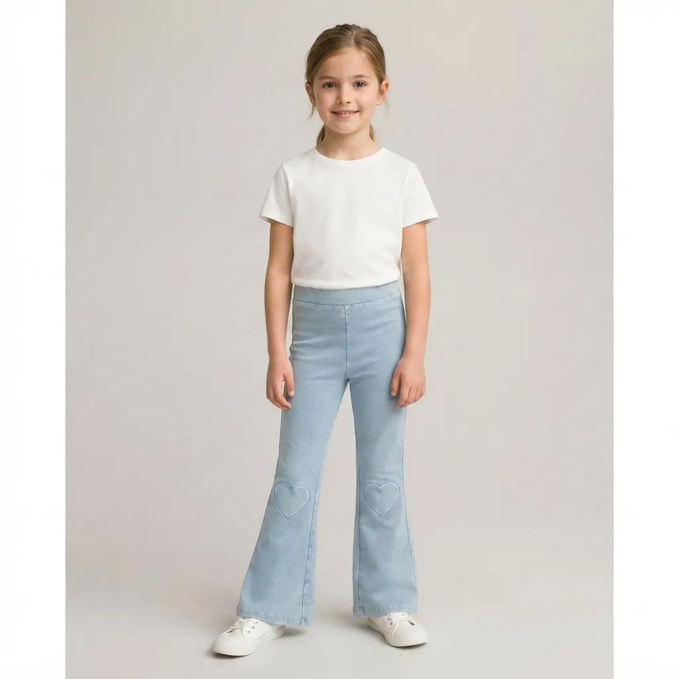 A young girl stands against a light gray studio background wearing a white short-sleeve t-shirt, light blue flared jeans with heart-shaped knee patches, and white sneakers.