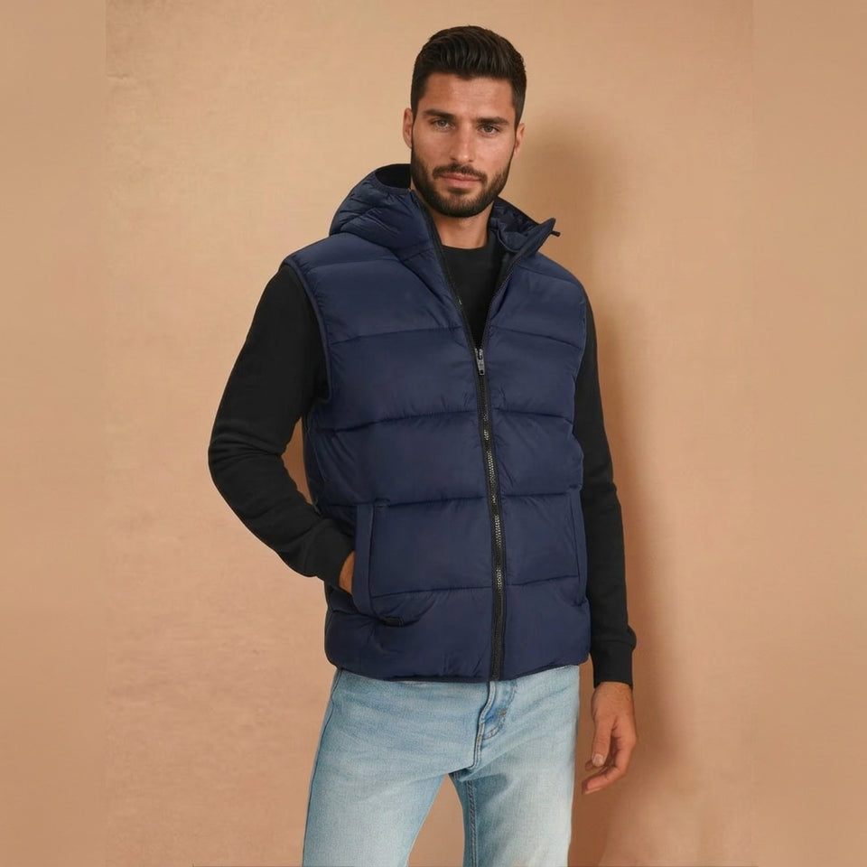 A man wearing a navy blue hooded puffer vest over a black long-sleeve shirt and light blue jeans, standing against a solid beige background.