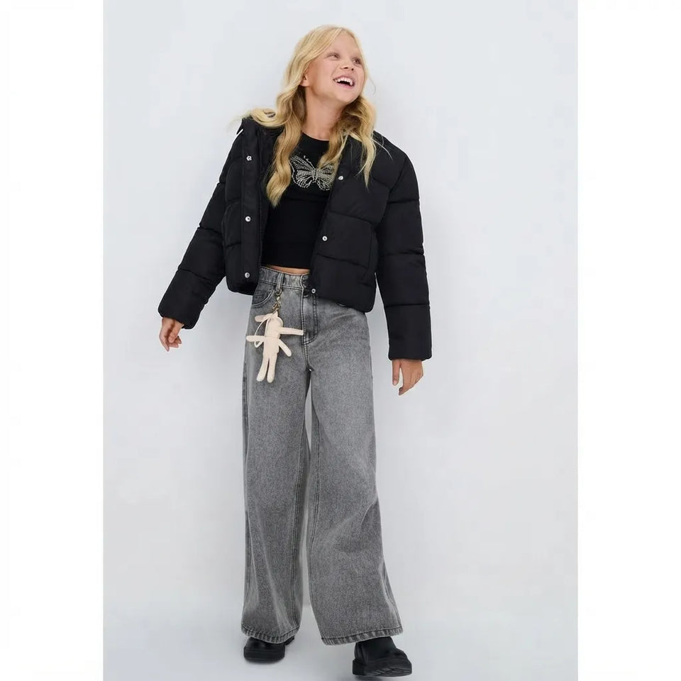 A woman modeling a black cropped puffer jacket over a black top, paired with wide-leg gray jeans featuring a beige dangling keychain, against a light gray studio background.