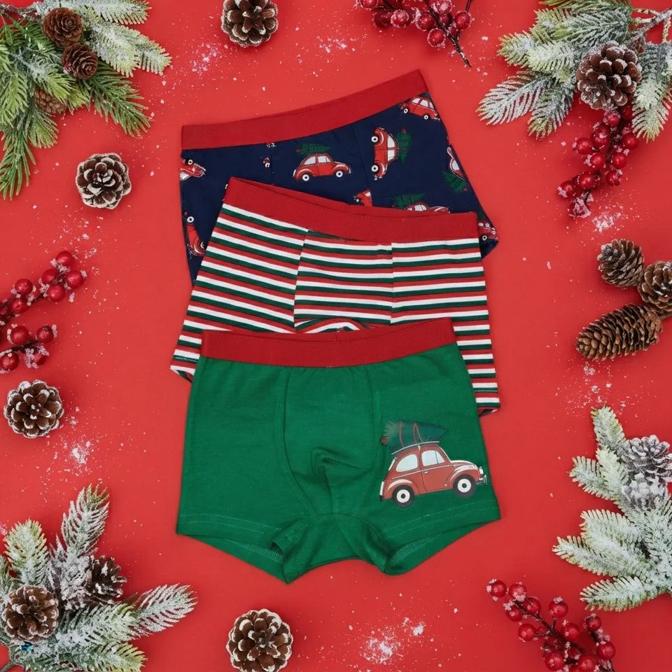 Three pairs of children's boxer briefs are arranged in a flat lay on a bright red background with holiday decorations. The top pair is navy blue with a red car print, the middle pair has red, white, and green stripes, and the bottom pair is green with a red car graphic.