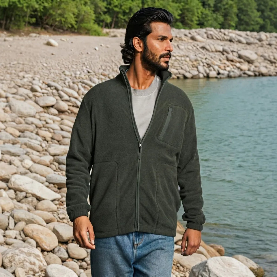 A man wearing an olive green zip-up fleece jacket, a gray t-shirt, and blue jeans stands on a rocky lakeshore.