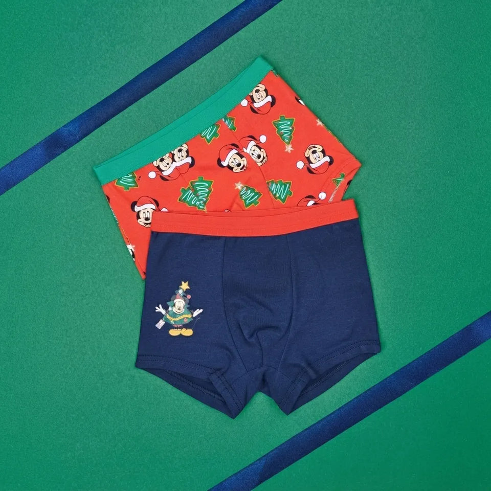 Three pairs of children's boxer briefs arranged in a stack on a green background with blue ribbon accents. The front pair is navy blue with a small Mickey Mouse Christmas tree graphic.