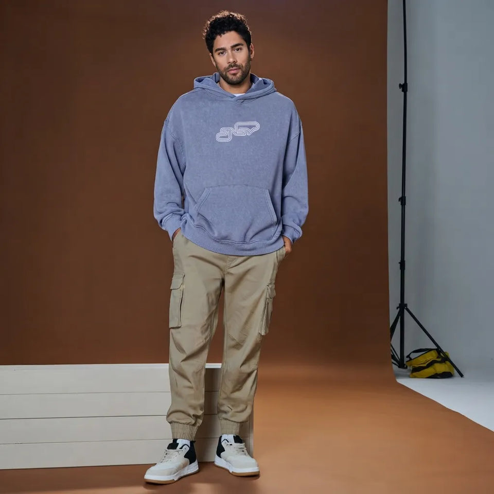 A man wearing a heathered periwinkle blue hoodie with a white chain logo on the chest, paired with beige cargo pants and white sneakers, standing in a studio setting with a brown backdrop.