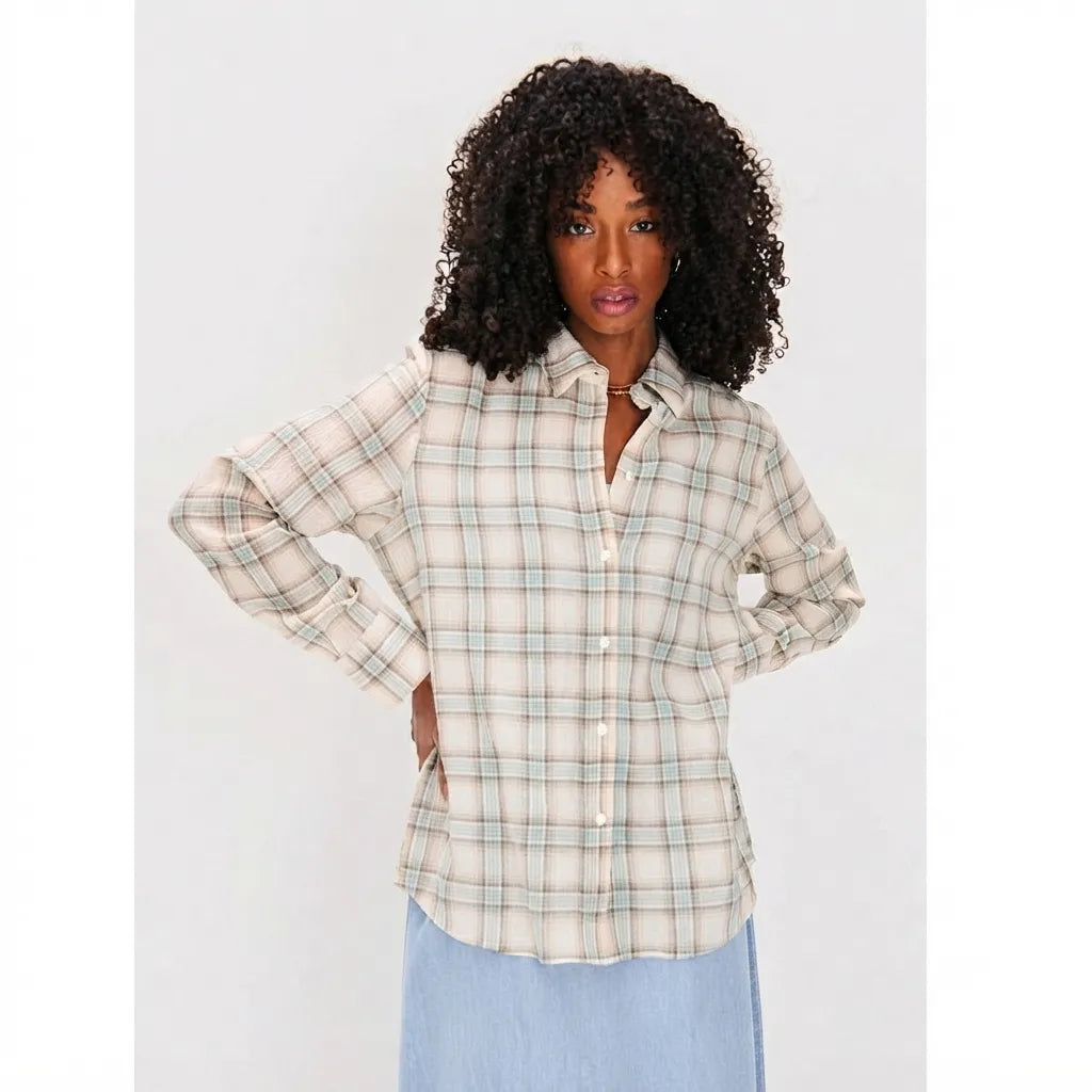 A woman with curly black hair wearing a cream-colored long-sleeve button-up plaid shirt with thin green and blue lines, paired with a light blue skirt, against a plain light gray background.