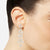 A close-up of a model's ear wearing a long, dangling crystal drop earring with a silver-toned floral design.